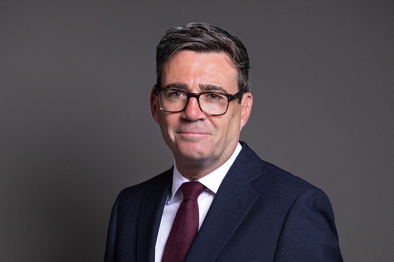 Official portrait of Prime Minister Andy Burnham wearing glasses and a dark suit with a maroon tie