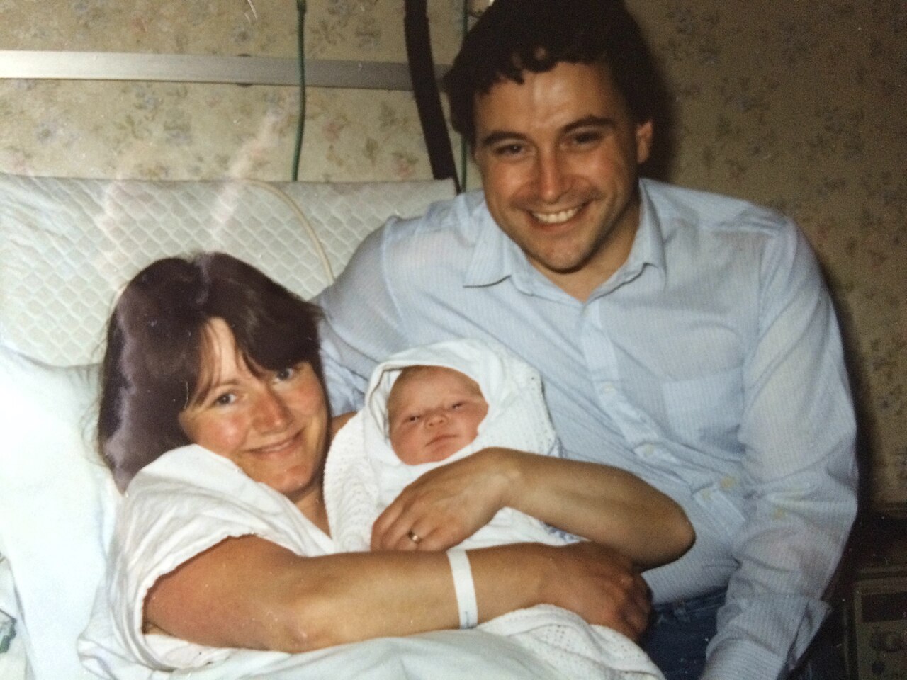 A new mother and father smile in a hospital room while holding their newborn baby, who is wrapped in a white knitted outfit.