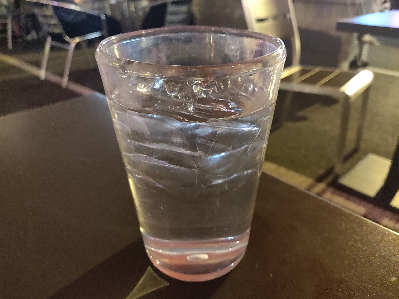 A glass of water with ice on a dark table, with empty metal chairs behind