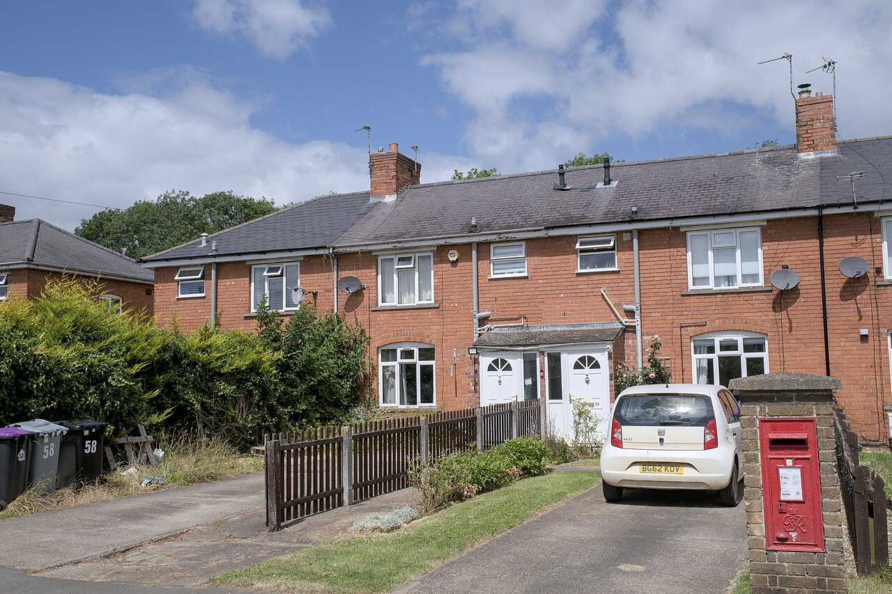 A terraced row of brick council houses on a residential UK street, with wheelie bins and parked cars outside.