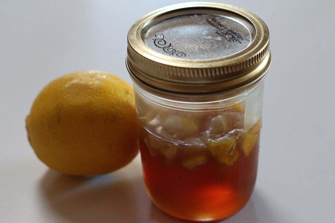 A jar of honey with lemon pieces beside a whole lemon