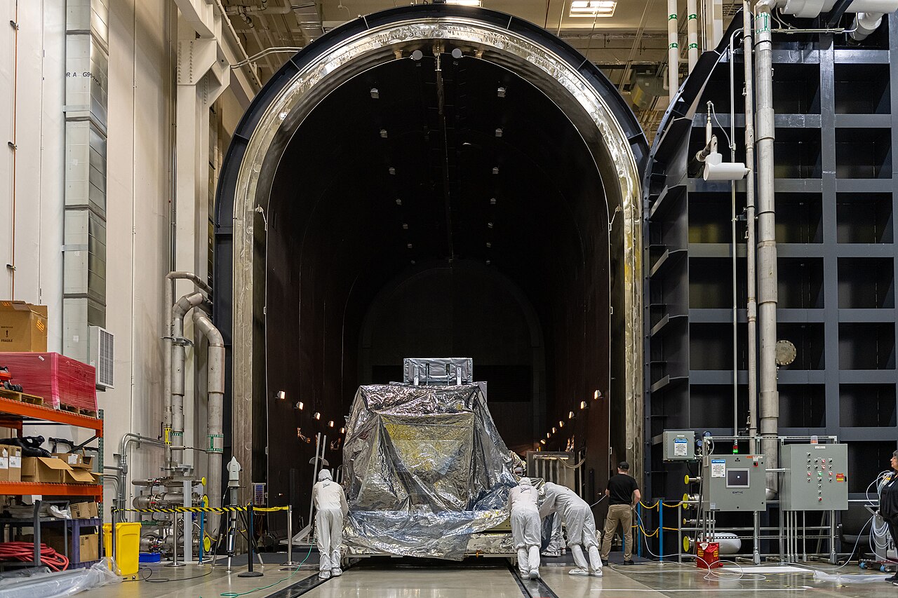 Workers, most in white protective suits, push the foil-wrapped Roman Wide Field Instrument towards a large testing chamber