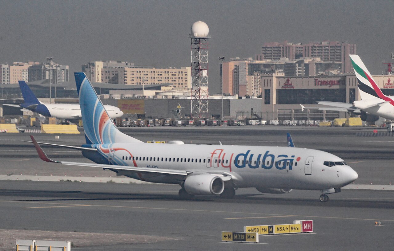 A flydubai Boeing 737 taxiing on an airport apron, with other aircraft and airport buildings behind