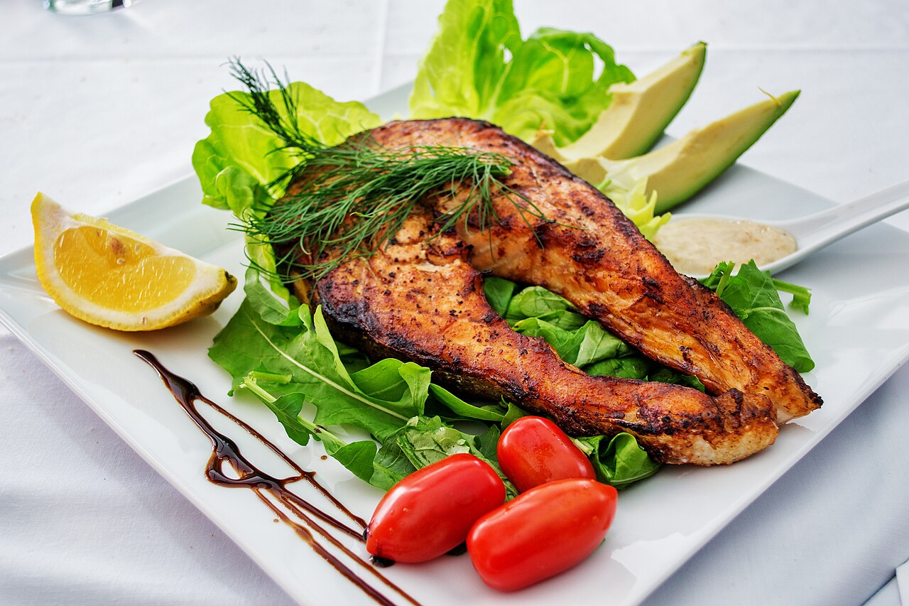 A grilled salmon fillet on a white plate with salad leaves, avocado, cherry tomatoes and a lemon wedge
