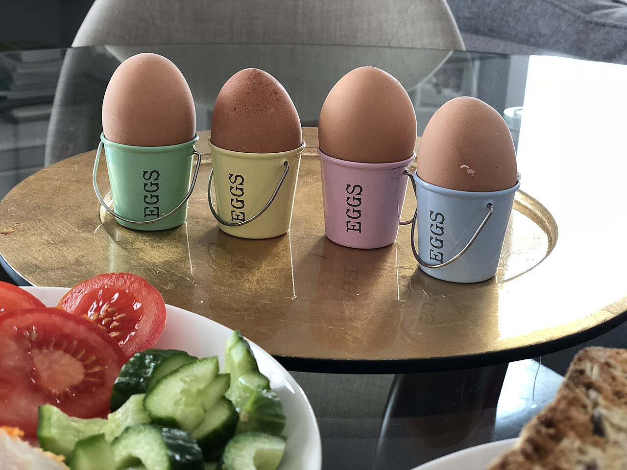 Four boiled eggs standing in small pastel egg cups on a gold-coloured table, with a plate of sliced tomato and cucumber in the foreground