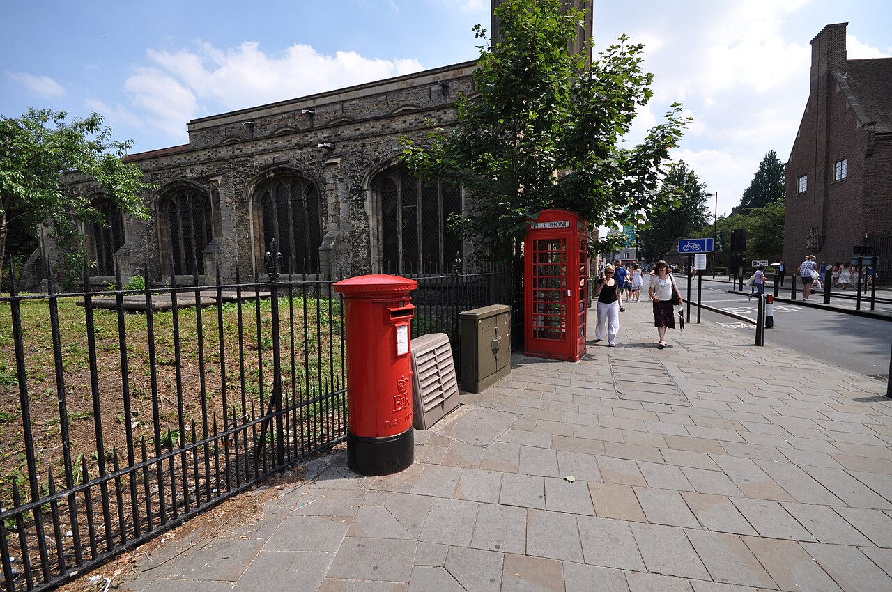 A red pillar box and a red telephone box on a paved street in front of a stone church in Cambridge