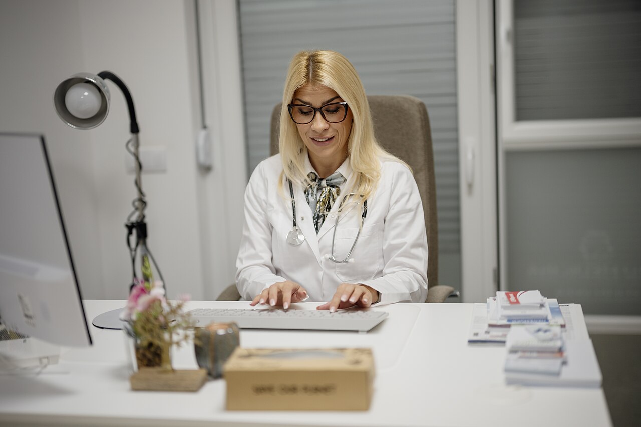 A doctor in a white coat typing at a desk in a consulting room