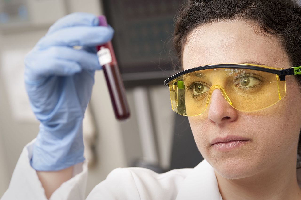 A laboratory worker in safety glasses and blue gloves holds up a tube of blood to examine it