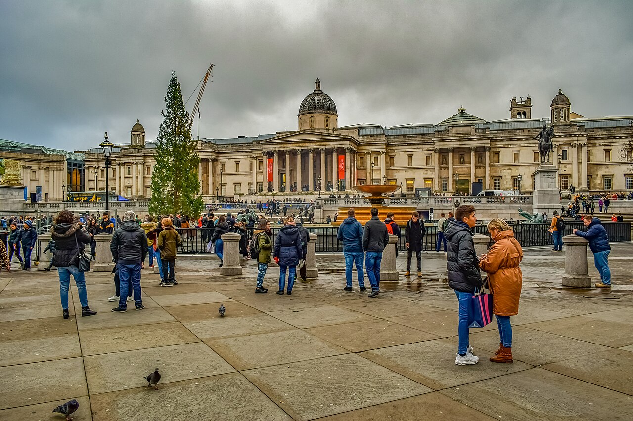 Visitors in Trafalgar Square in front of the National Gallery, with a tall Christmas tree on the left under a grey sky