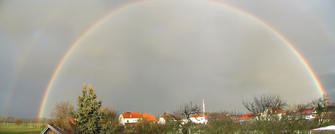 A full double rainbow arches over a village with orange-roofed houses under a grey sky, a fainter second bow just visible outside the first