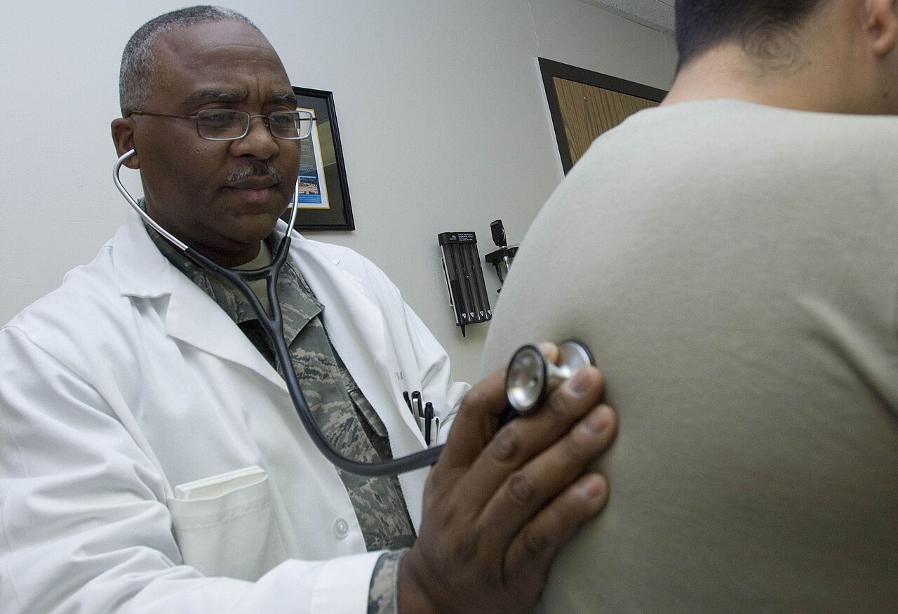 A doctor in a white coat listens through a stethoscope to the breathing of a patient seen from behind in a consulting room