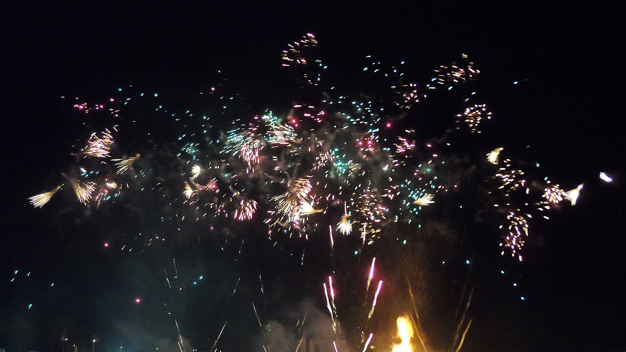 Colourful fireworks bursting in a night sky above a glowing bonfire flame at the Lewes Bonfire Night display, East Sussex