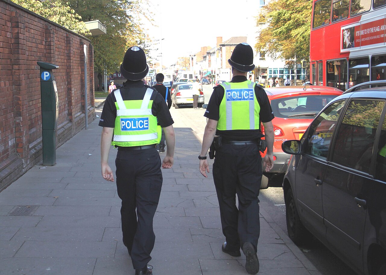 Two police officers walking on foot patrol along an English high street, seen from behind, with POLICE on their high-visibility vests.