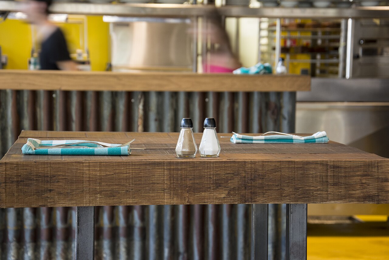 Two glass shakers with black lids on a wooden restaurant table, between two folded striped napkins