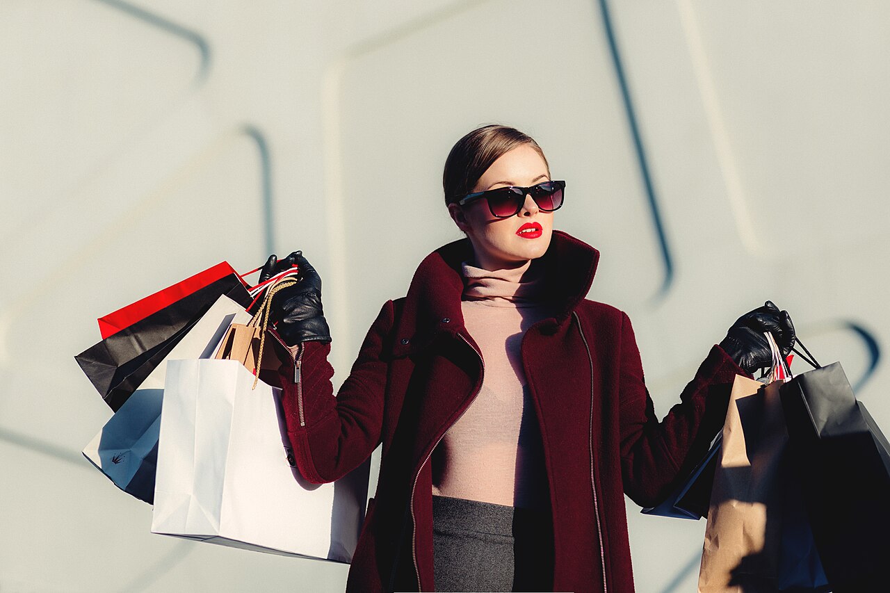 A woman in a burgundy coat and sunglasses holds several paper shopping bags against a pale wall.