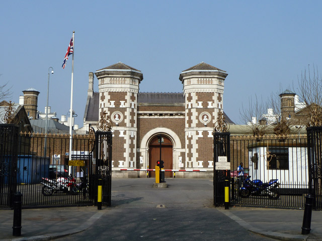 The brick gatehouse and iron entrance gates of Wormwood Scrubs prison in London, with a Union flag flying above the gate towers