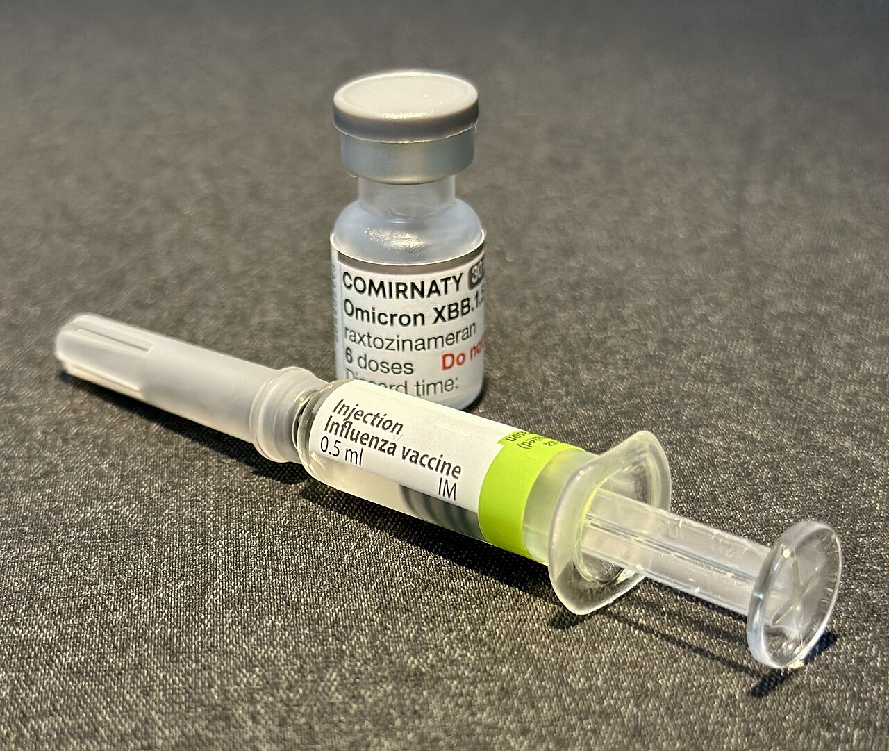 A prefilled influenza vaccine syringe lying in front of a vial of COVID-19 vaccine on a grey surface