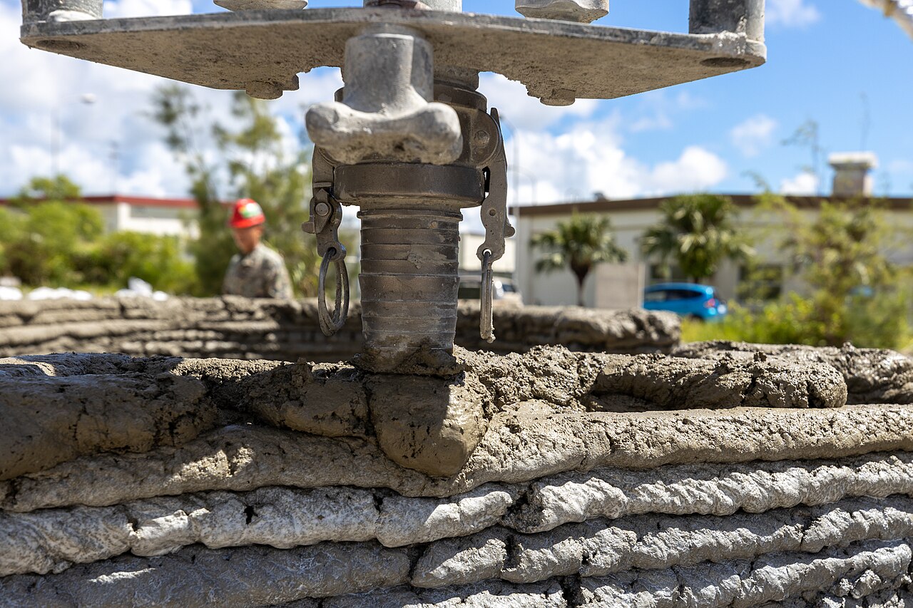 A 3D concrete printer nozzle depositing a fresh layer of wet concrete on a wall built up in rough horizontal layers