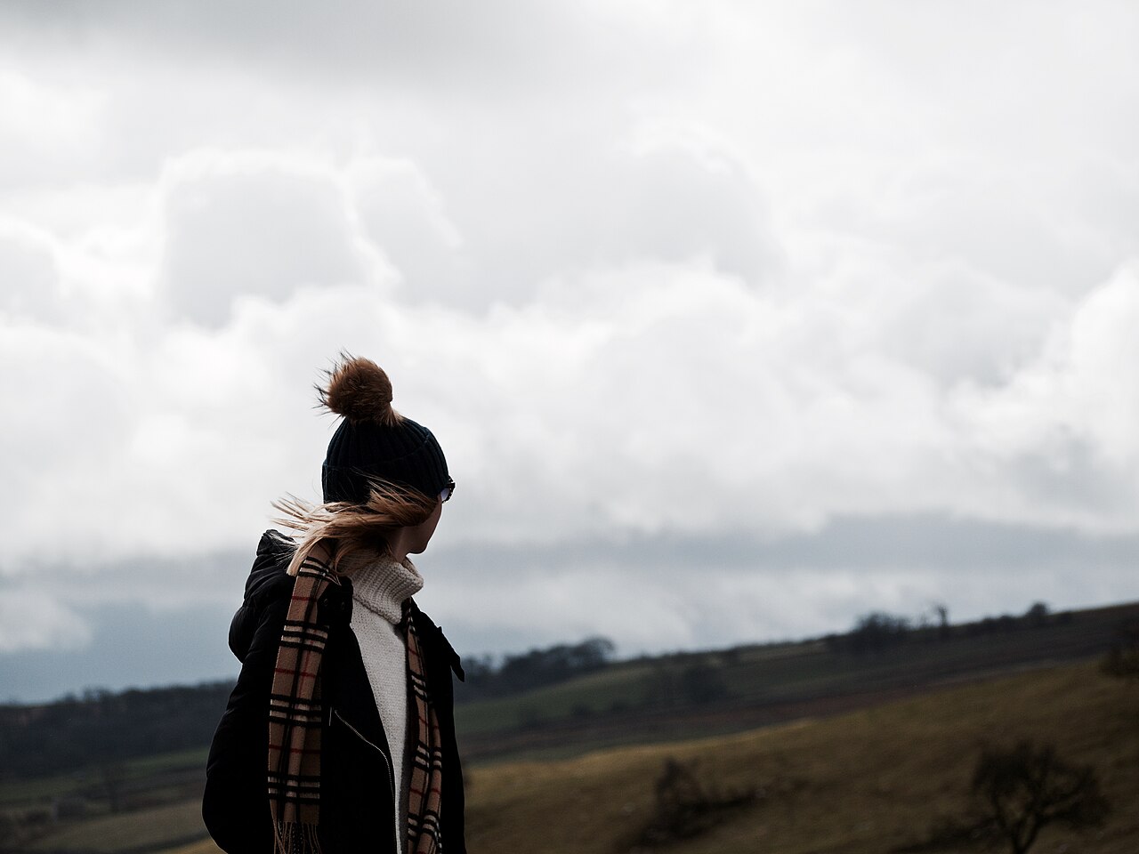 A woman in a woolly hat and checked scarf looks away across a windswept hillside under heavy grey cloud