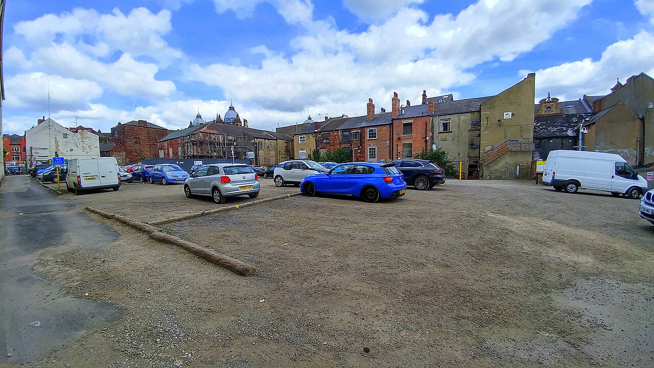 Cars and vans parked on an open gravel car park in Leeds, with brick buildings and a cloudy sky behind