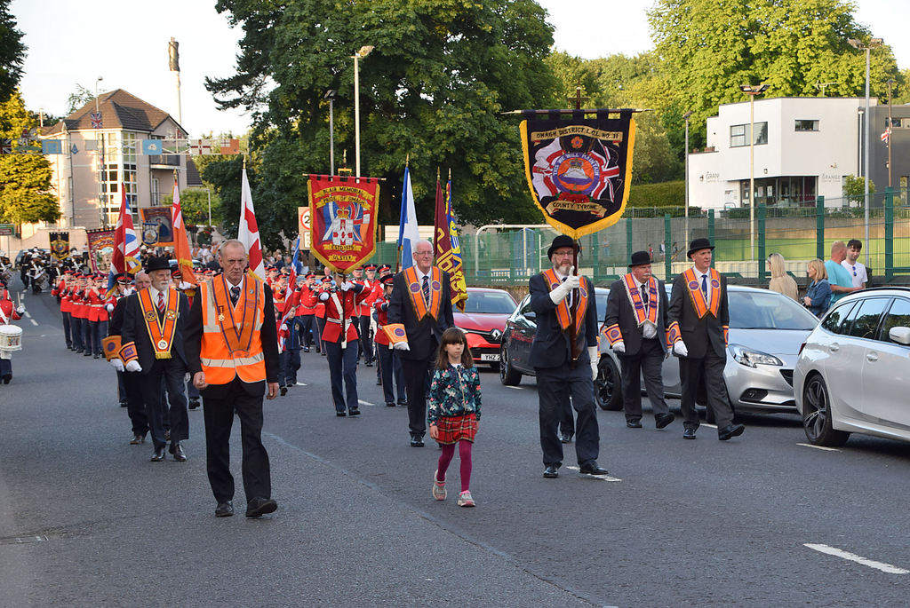 Orangemen wearing orange sashes and carrying lodge banners during a parade through a Northern Ireland town, led by a marching band
