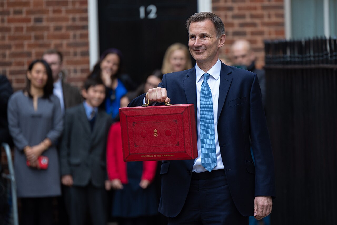 A Chancellor of the Exchequer holds the red Budget box outside Downing Street on an earlier Budget day, with onlookers behind.