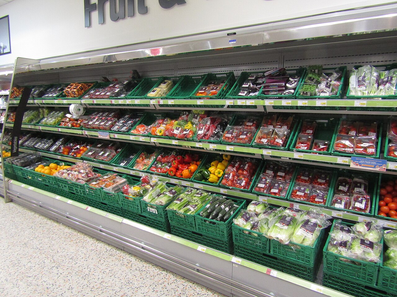 A supermarket chilled display of fruit and vegetables in green crates, with peppers, tomatoes and salad in packs and price labels along the shelf edges.