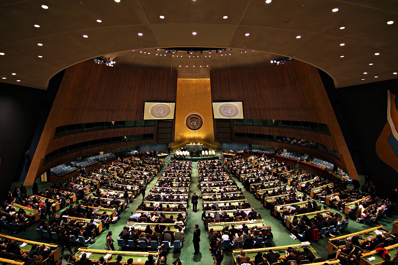 Delegates seated in the UN General Assembly hall in New York, with the rostrum and the UN emblem at the back