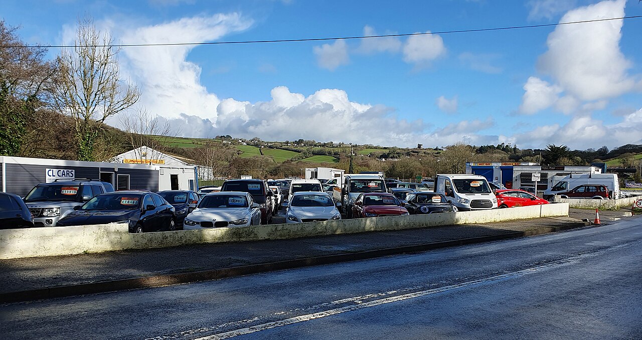 Used cars parked on a dealer’s forecourt beside a road in Cornwall, with price labels on the windscreens
