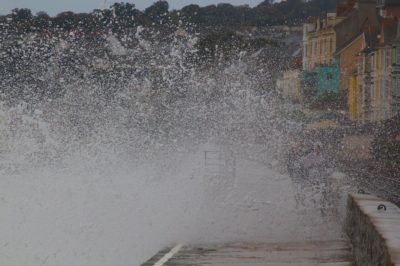 Waves breaking over the sea wall at Dawlish on the Devon coast, with spray rising above the railway and seafront houses