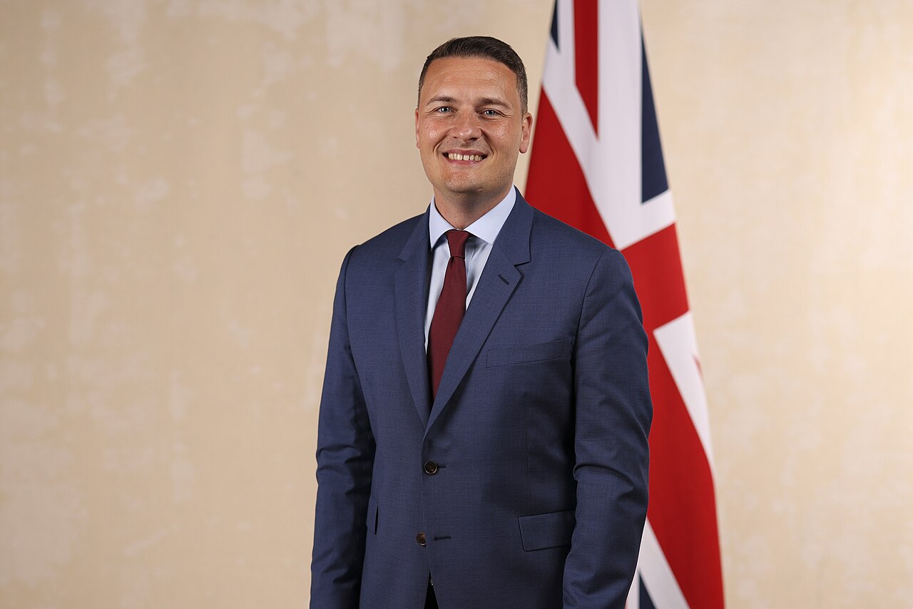 Official portrait of Wes Streeting in a blue suit and red tie, standing beside a Union Jack flag