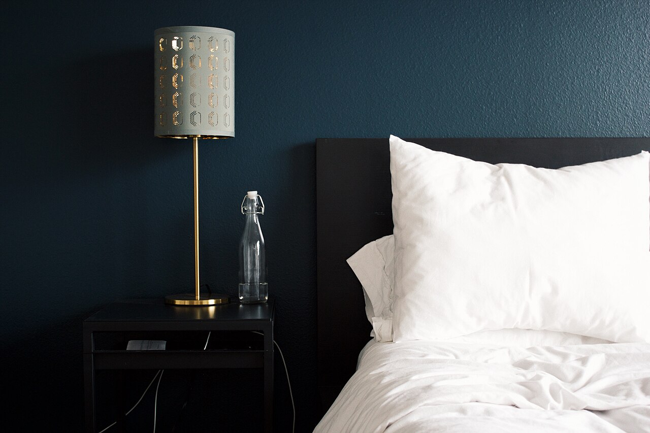 A bedside table with a lamp and a glass bottle of water beside a bed with white pillows and sheets against a dark blue wall