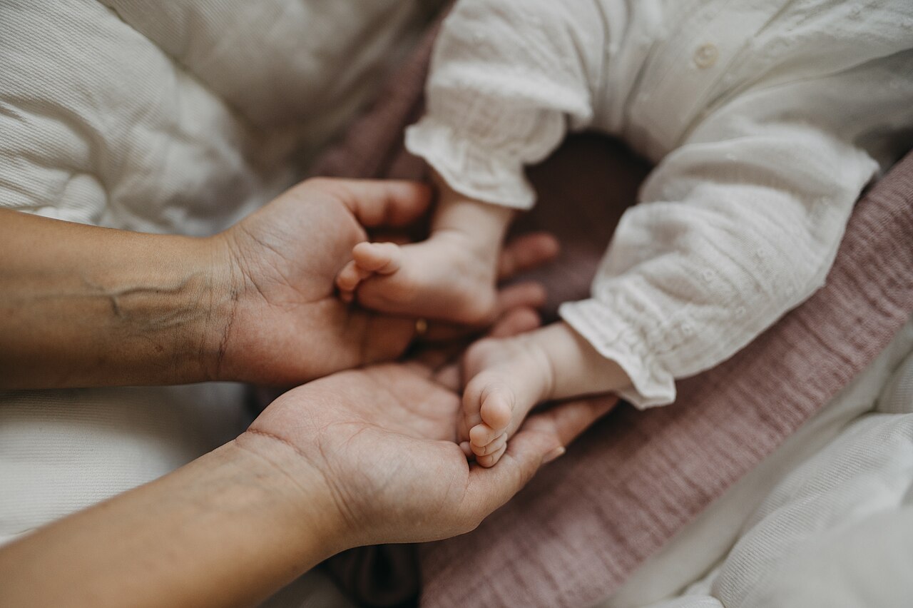 Two adult hands cradling the feet of a baby wrapped in a white cotton romper on a soft pink blanket
