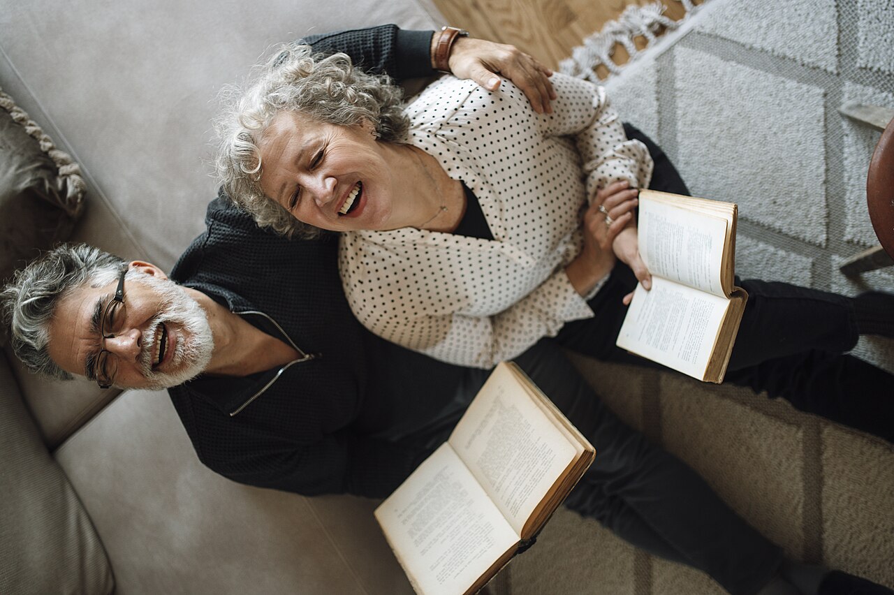 An older man and woman laughing together at home, each holding an open book