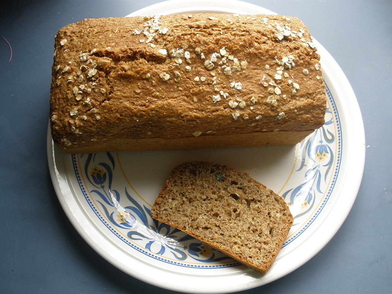 A loaf of wholemeal brown bread topped with oats, with one slice cut to show its wholegrain texture