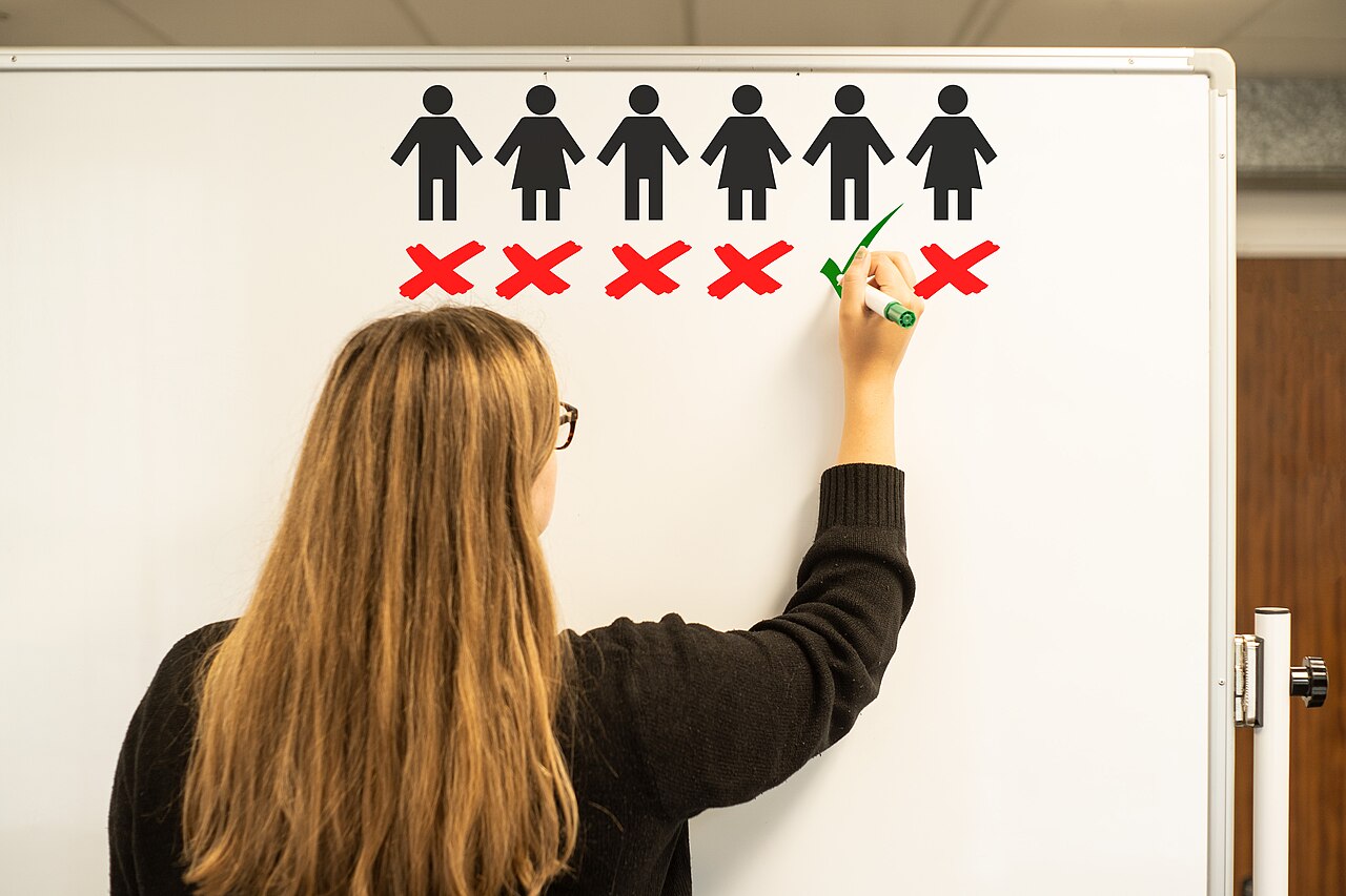 A woman with her back to the camera ticks one figure and crosses out five others on a whiteboard