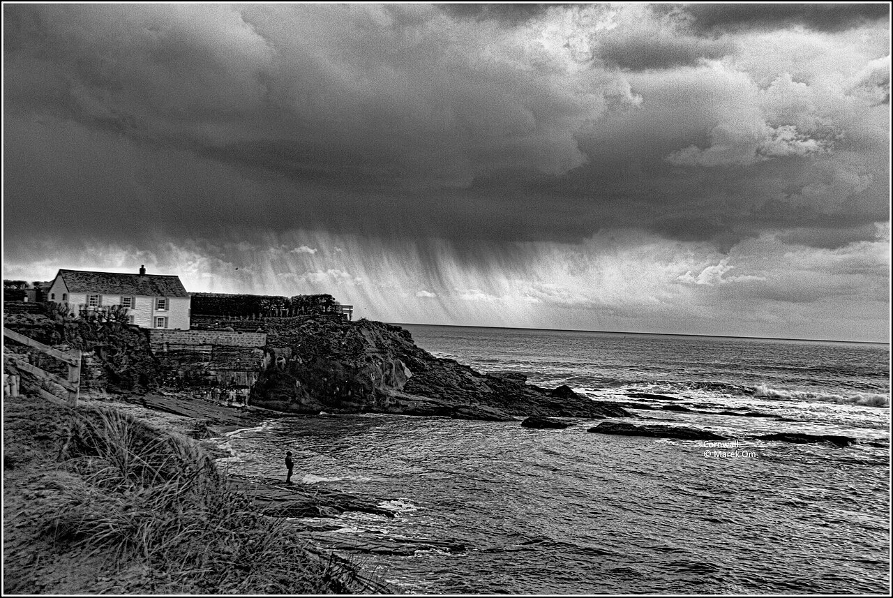 Black-and-white photograph of rain falling from dark clouds over a rocky stretch of Cornish coast, with a house on the cliff and a lone figure at the water’s edge