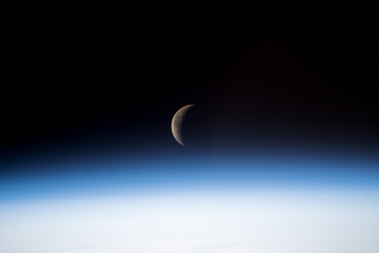 A waning crescent Moon above the blue glow of Earth’s atmosphere, photographed from the International Space Station