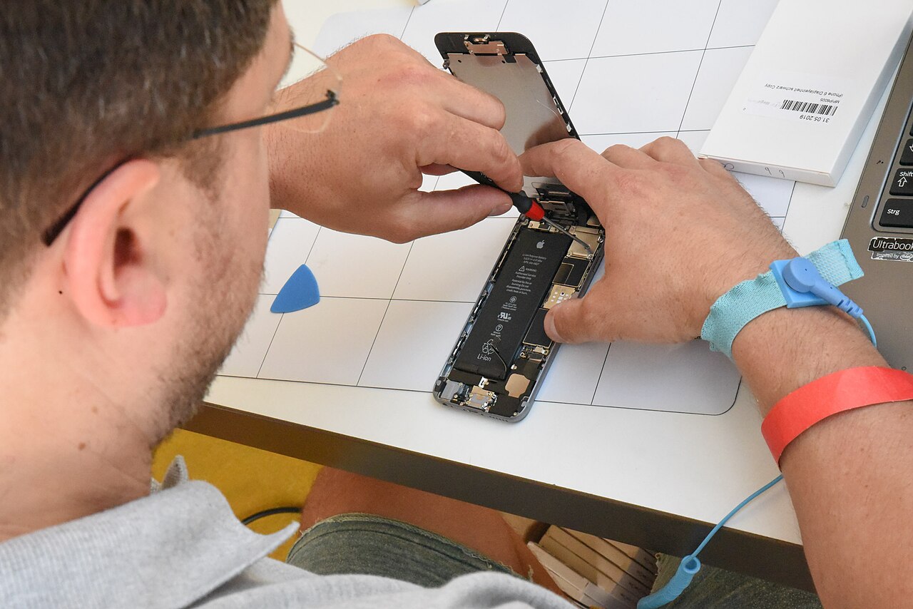 A man wearing an anti-static wrist strap uses a screwdriver to open a smartphone at a repair workshop table