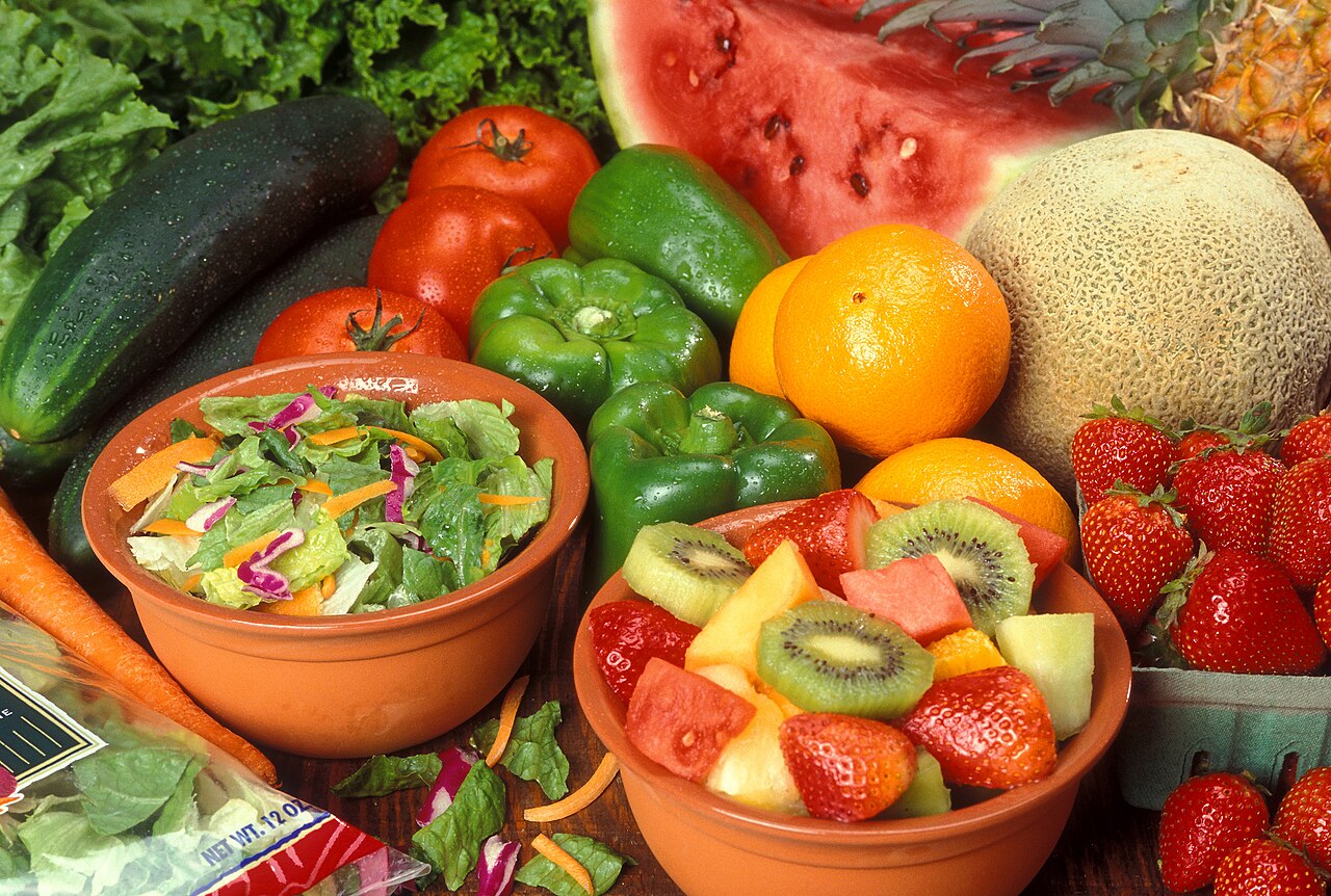Fresh fruit and vegetables including tomatoes, peppers, oranges, melon, strawberries and two bowls of salad and chopped fruit