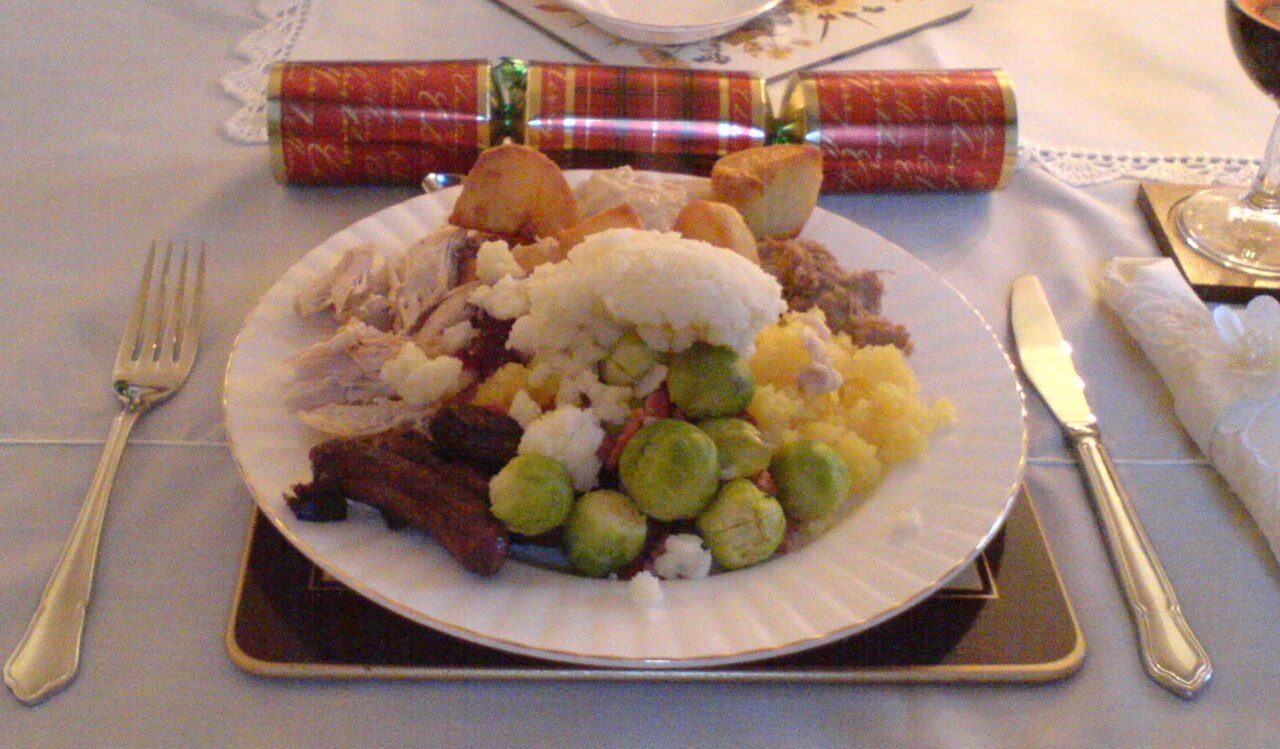 A plate piled with roast meat, roast potatoes, sprouts, cauliflower and sausages beside a Christmas cracker and a glass of red wine