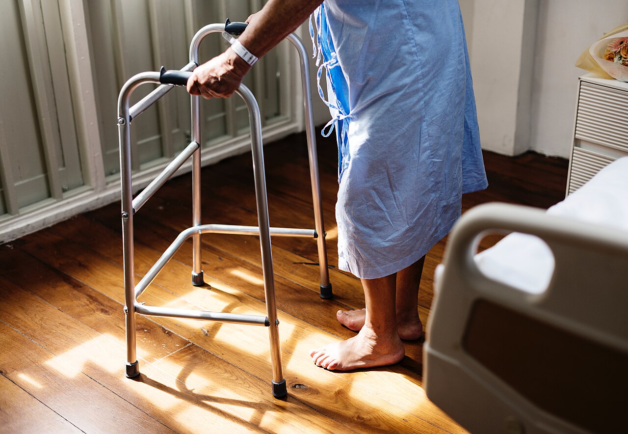 A person using a metal walking frame to stand, photographed from behind near a hospital-style bed.