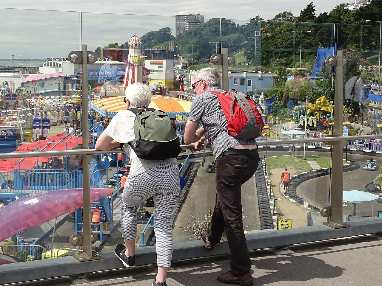 An older couple with rucksacks lean on a glass rail overlooking a seaside amusement park in Southend-on-Sea, Essex